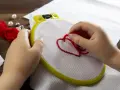 Mani di bambino creano un cuore rosso con il punto erba a mano su tela bianca, circondato da perle e gomitoli di lana.