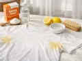 Maglietta bianca con macchie gialle sotto le ascelle, accanto a bicarbonato, aceto, limoni e spazzola per bucato.