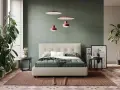 Letto moderno con testiera capitonné, copriletto verde e cuscini bianchi. Parti del letto ben visibili, con comodini e piante.
