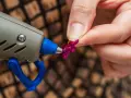Applicazione strass su tessuto con colla a caldo. Una mano tiene un fiorellino fucsia mentre la pistola a caldo è pronta per incollare.