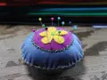 Puntaspilli fai da te a forma di fiore, con petali gialli su sfondo viola e base blu, decorato con cuciture a zig-zag verdi e gialle.