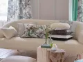 Divano in tessuto bouclé color crema, pronto per essere rifoderato, con cuscini decorativi e tende floreali sullo sfondo.