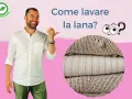 Uomo sorridente indica come lavare lana a mano. Pile di maglioni in primo piano.