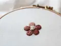 Piccolo fiore ricamato a punto pieno su tela grezza, con petali marroni e centro bianco.