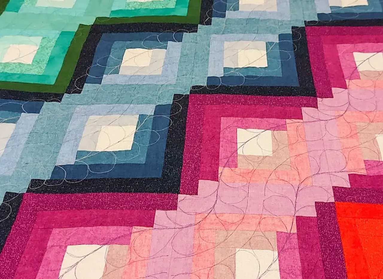 Dettaglio di una coperta realizzata con la tecnica patchwork, con blocchi a gradiente di colore dal blu al rosa, rifinita con cuciture decorative.