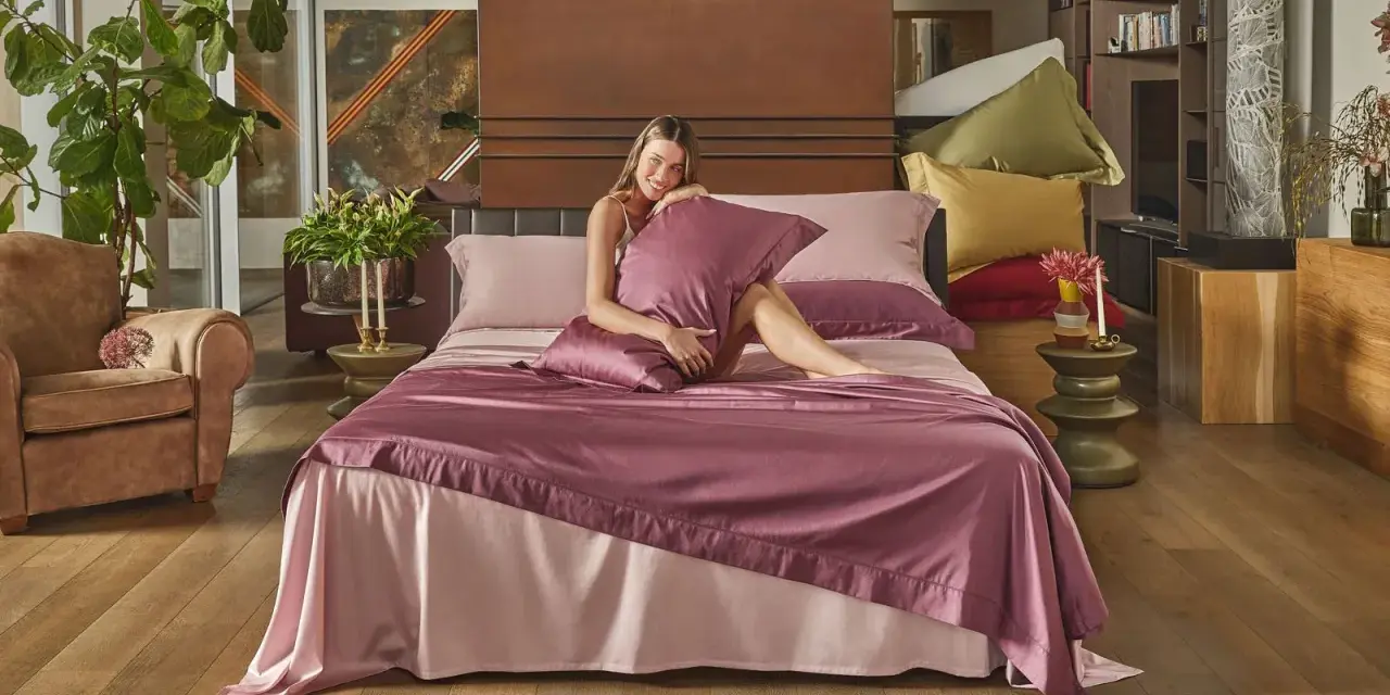 Donna sorridente su un letto con lenzuola rosa e copriletto prugna, che illustra la differenza tra piumone e trapunta.