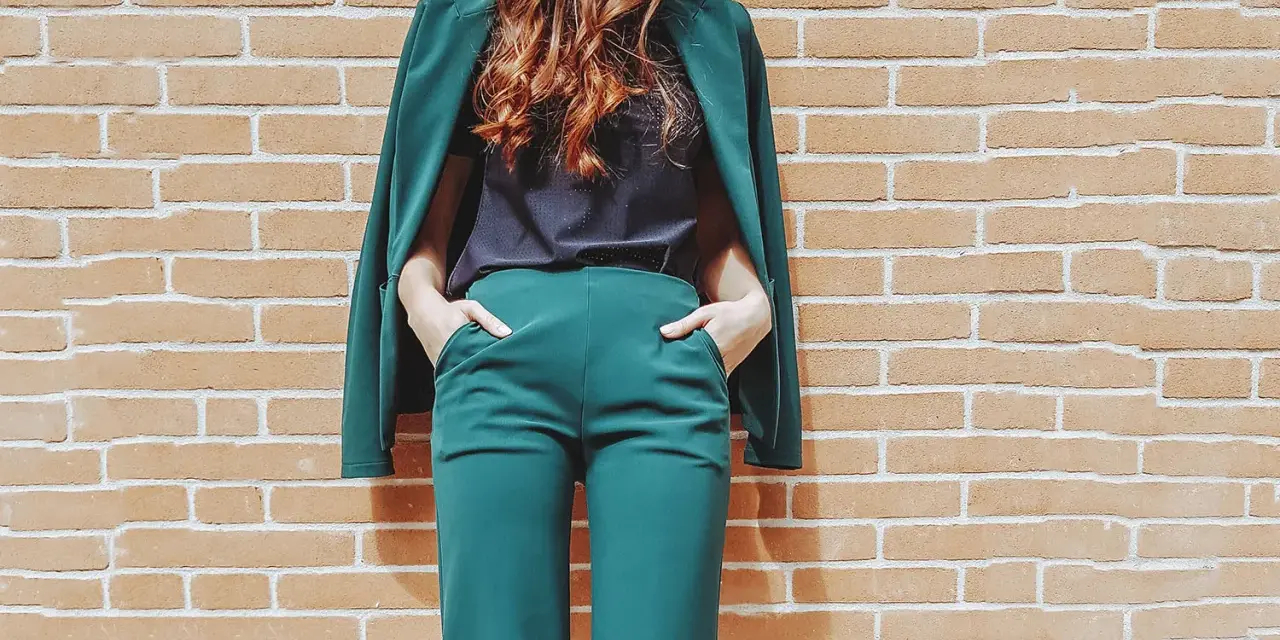 Donna con tailleur verde smeraldo, composto da giacca e pantaloni a palazzo, con le mani in tasca, appoggiata a un muro di mattoni.