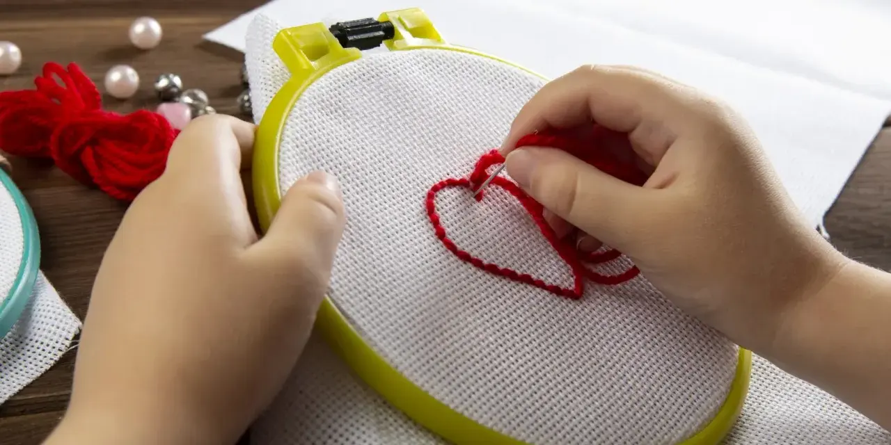 Mani di bambino creano un cuore rosso con il punto erba a mano su tela bianca, circondato da perle e gomitoli di lana.