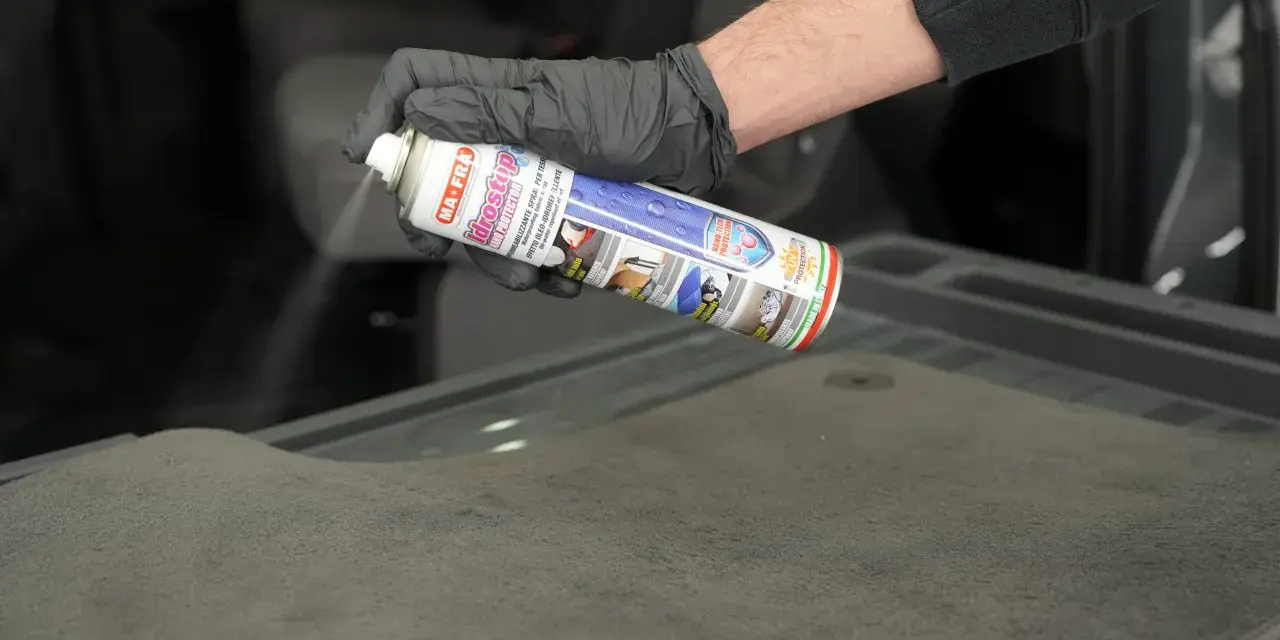 Mano con guanto nero spruzza spray Idrostop su tessuto grigio per impermeabilizzare.