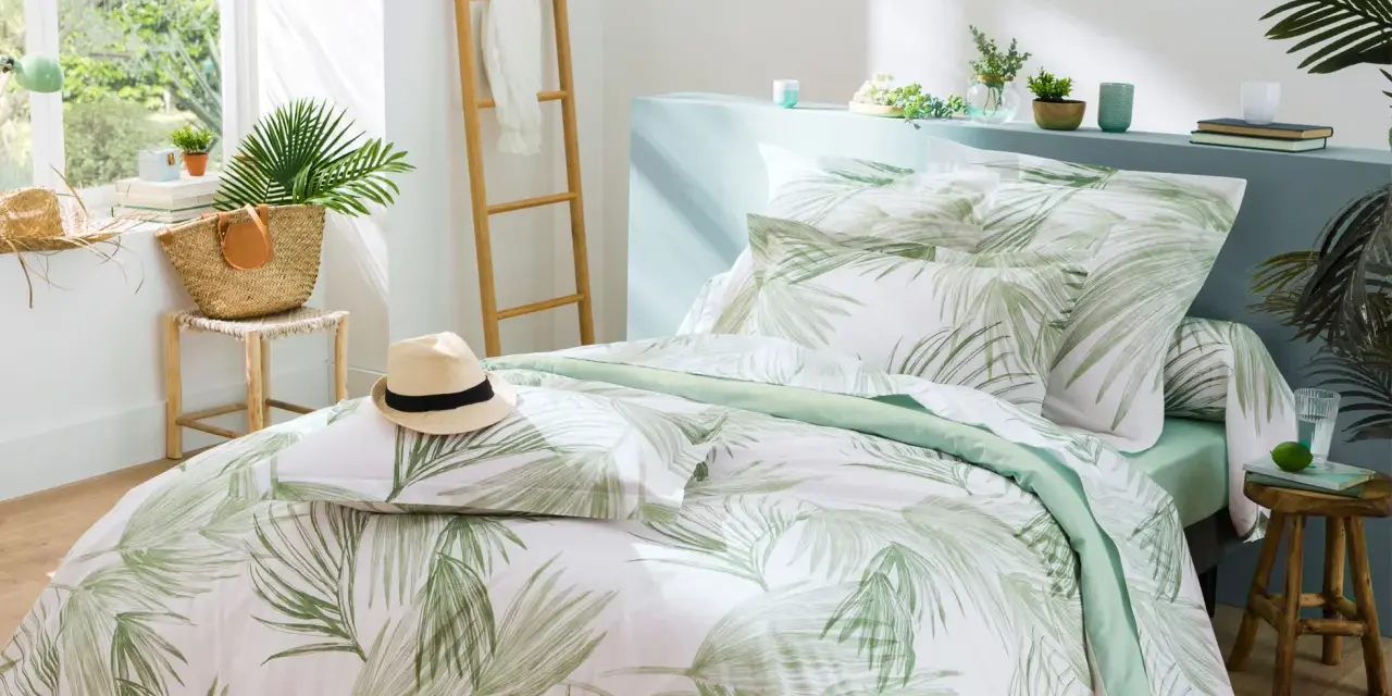 Un letto preparato per l'estate con biancheria a tema tropicale, un cappello di paglia e una borsa di vimini.