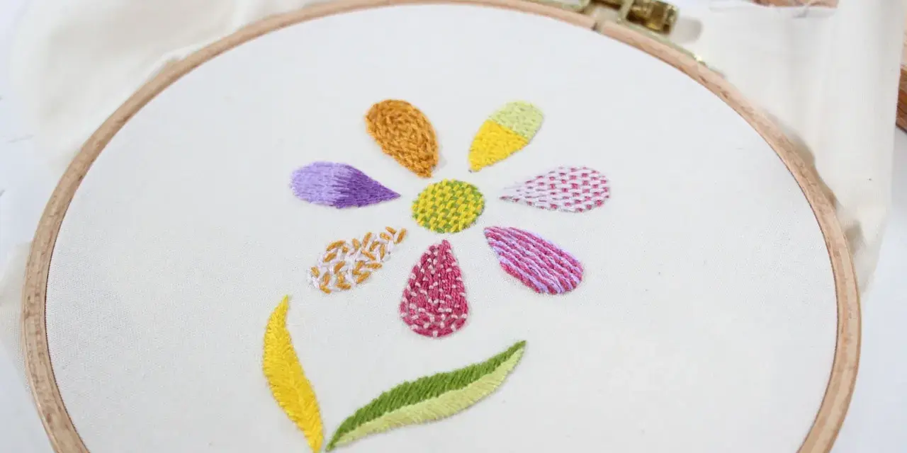 Fiore ricamato su tela bianca con petali colorati e diversi punti ricamo per riempire, che mostrano varie texture e tecniche.