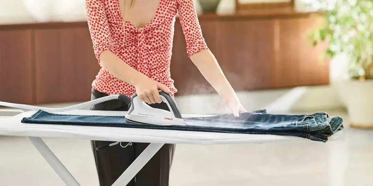 Donna stira un paio di jeans su un asse da stiro, usando un ferro da stiro a vapore. Impara come stendere i pantaloni per un look impeccabile.