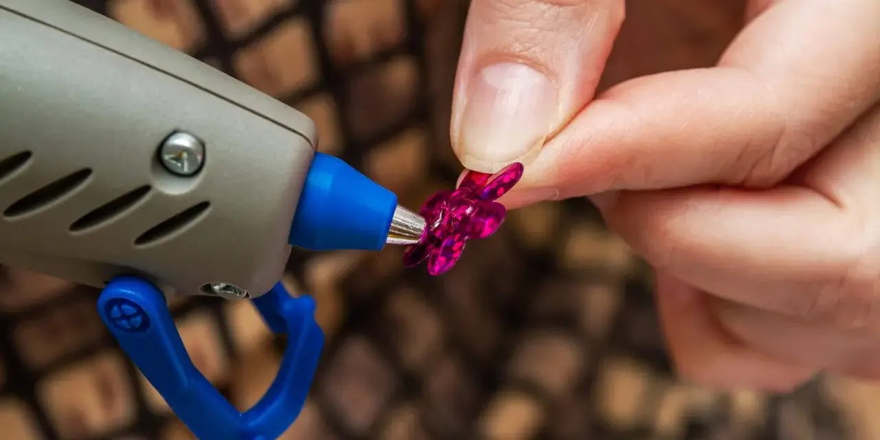 Applicazione strass su tessuto con colla a caldo. Una mano tiene un fiorellino fucsia mentre la pistola a caldo è pronta per incollare.