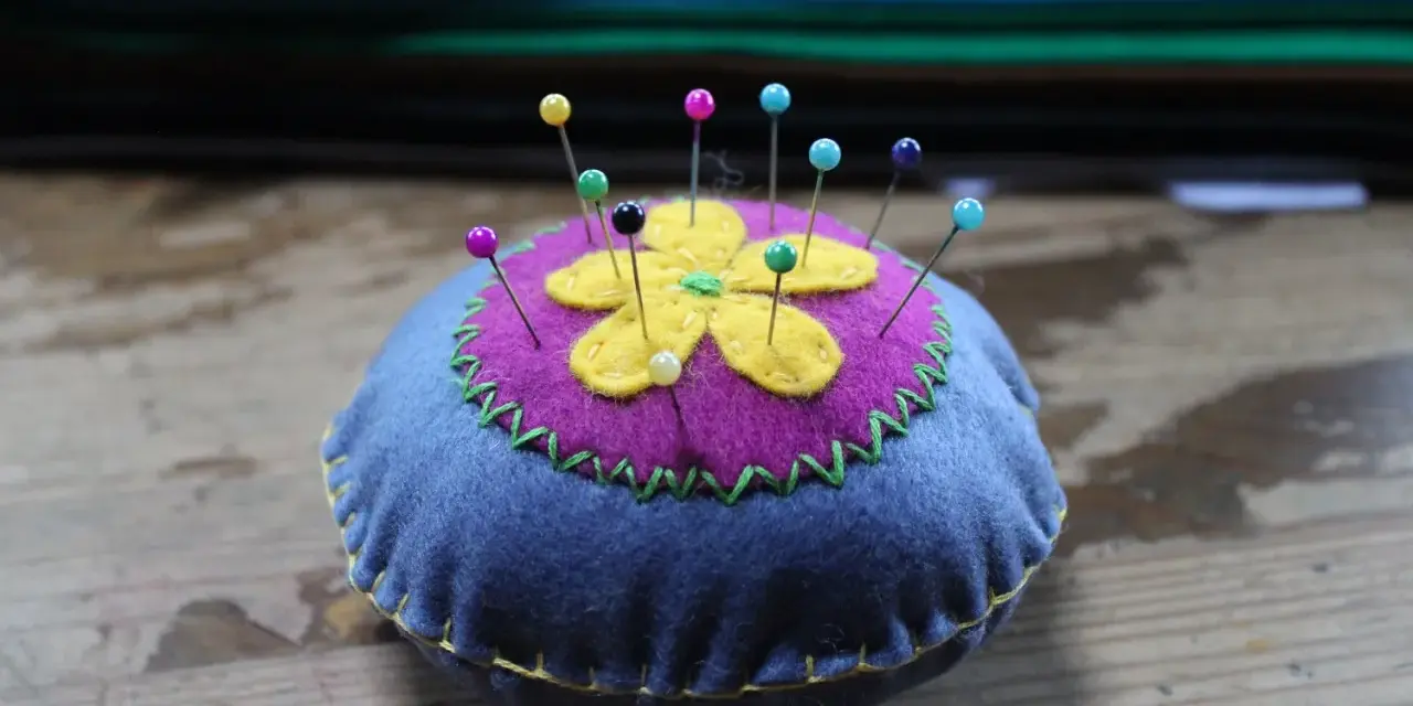 Puntaspilli fai da te a forma di fiore, con petali gialli su sfondo viola e base blu, decorato con cuciture a zig-zag verdi e gialle.