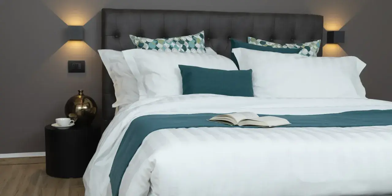 Letto con trapunta bianca a righe, cuscini coordinati e un libro aperto. Ecco come mettere la trapunta nel letto per un riposo perfetto.