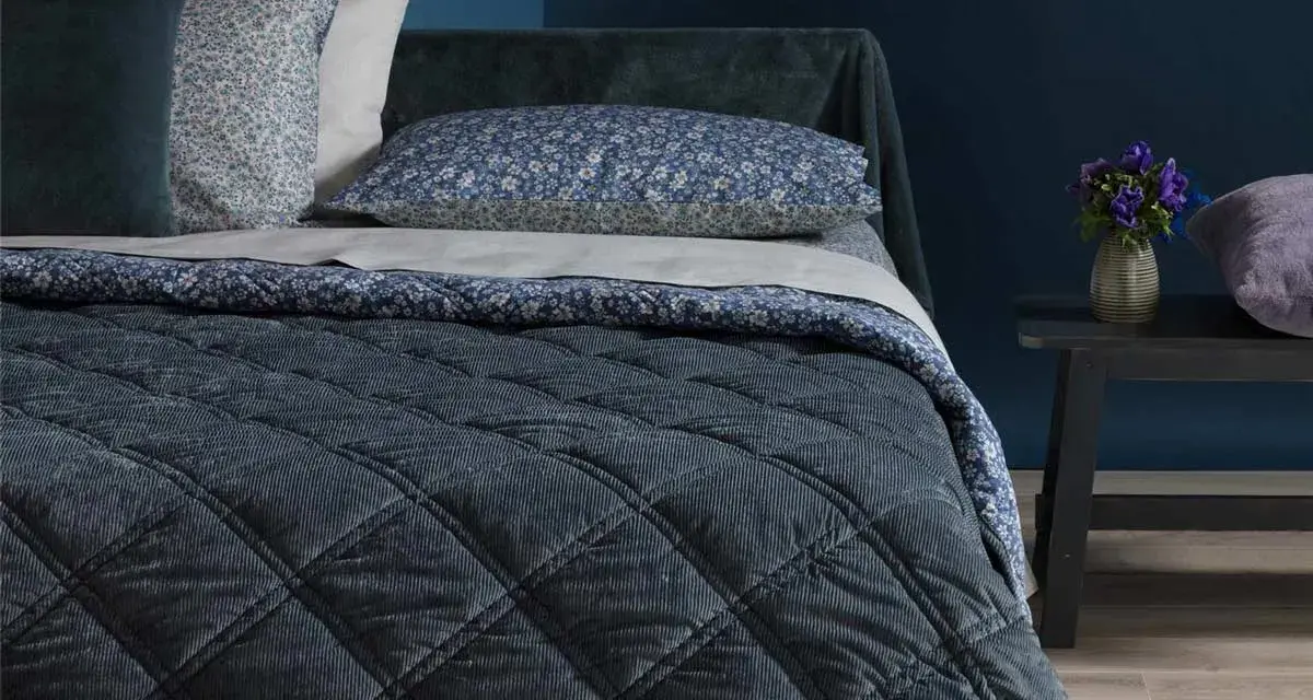Letto con copriletto trapuntato blu scuro, cuscini floreali e un dettaglio che ricorda che il velluto si rovina con acqua.