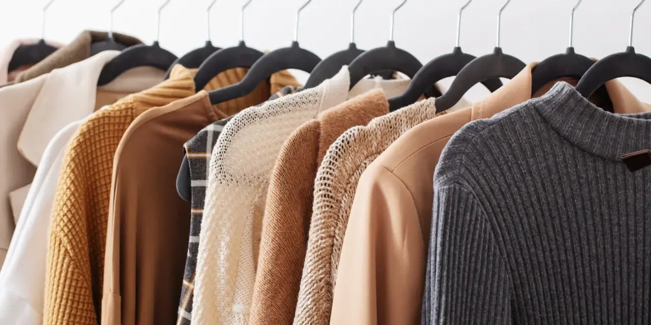 Una fila di capi d'abbigliamento in tonalità neutre, tra cui un maglione lavorato a maglia e un blazer in tessuto cupro lavaggio, pronti per essere indossati.