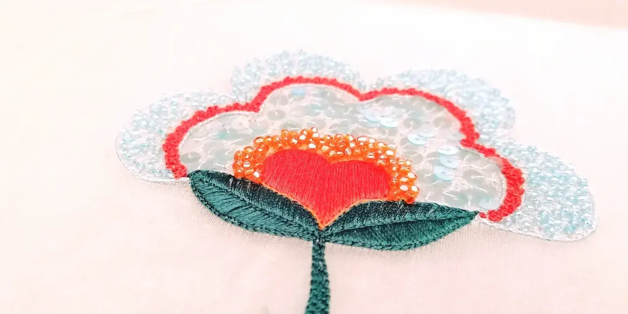 Un fiore stilizzato con petali azzurri e paillettes, un cuore rosso al centro e perline arancioni. Un'opera di ricamo francese che cattura la luce.