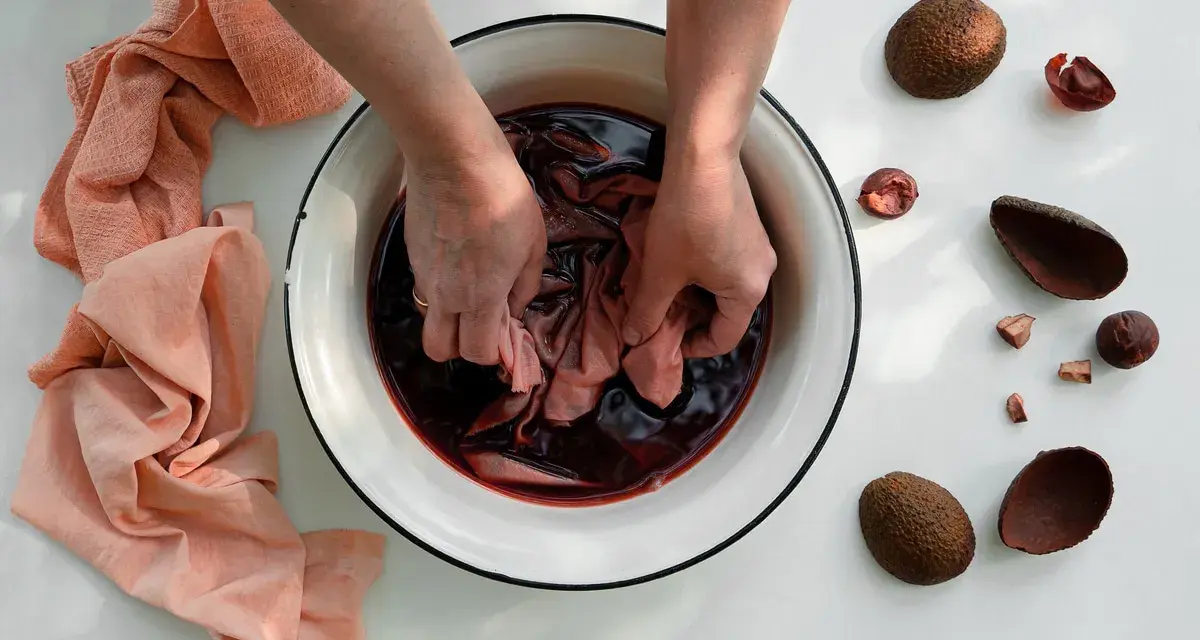 Mani che tingono un tessuto in una bacinella con un colorante naturale per tessuti. Avocado e bucce intorno.