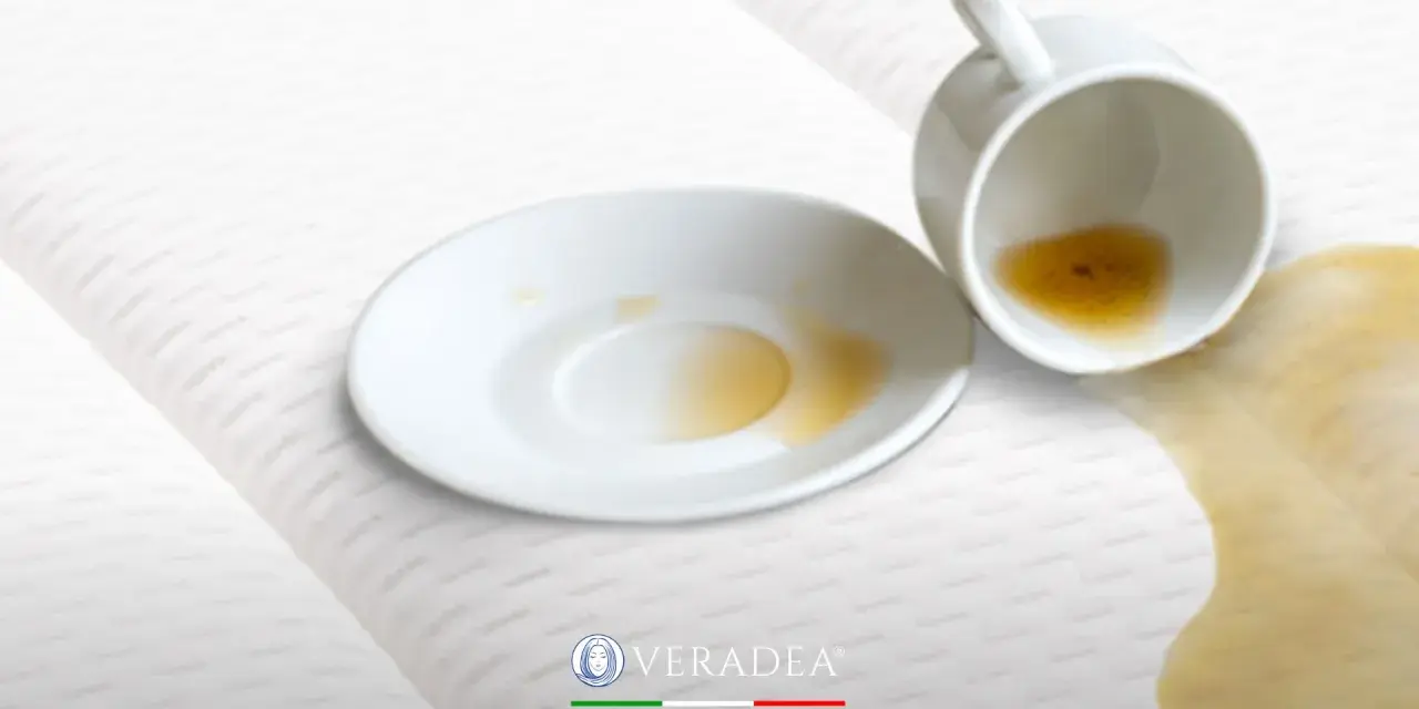 Tazza rovesciata su un materasso bianco, con liquido marrone versato. Scopri come disinfettare il materasso per eliminare macchie e odori.