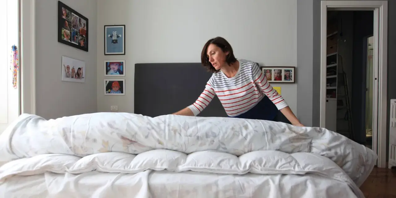 Donna con maglia a righe prova a mettere il copripiumino su un letto disfatto.