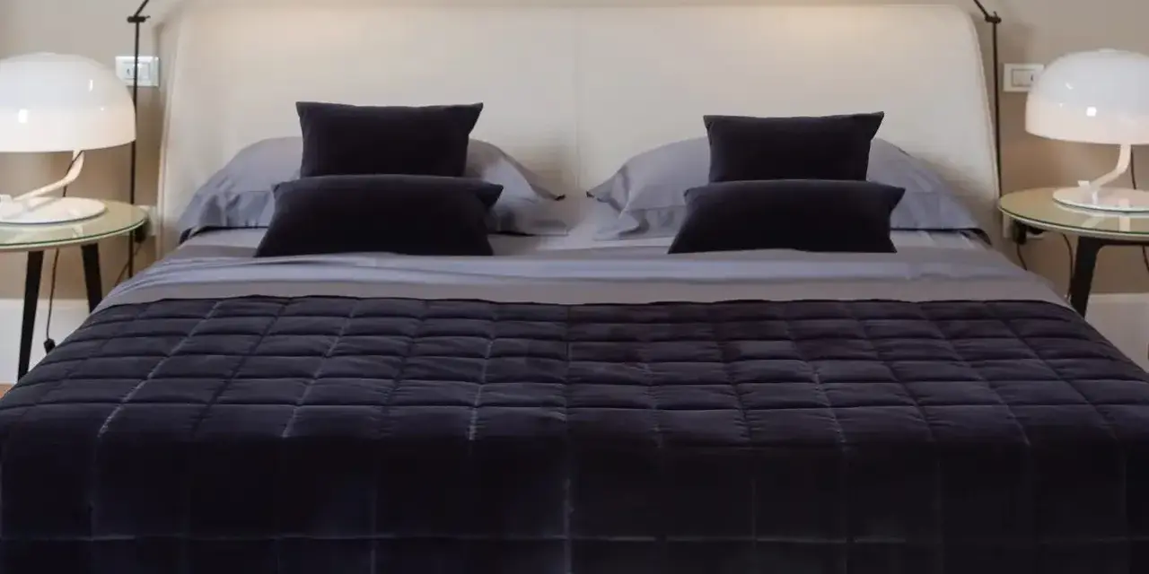 Un letto impeccabile, con copriletto trapuntato scuro e cuscini coordinati, mostra come rifare il letto in modo elegante.
