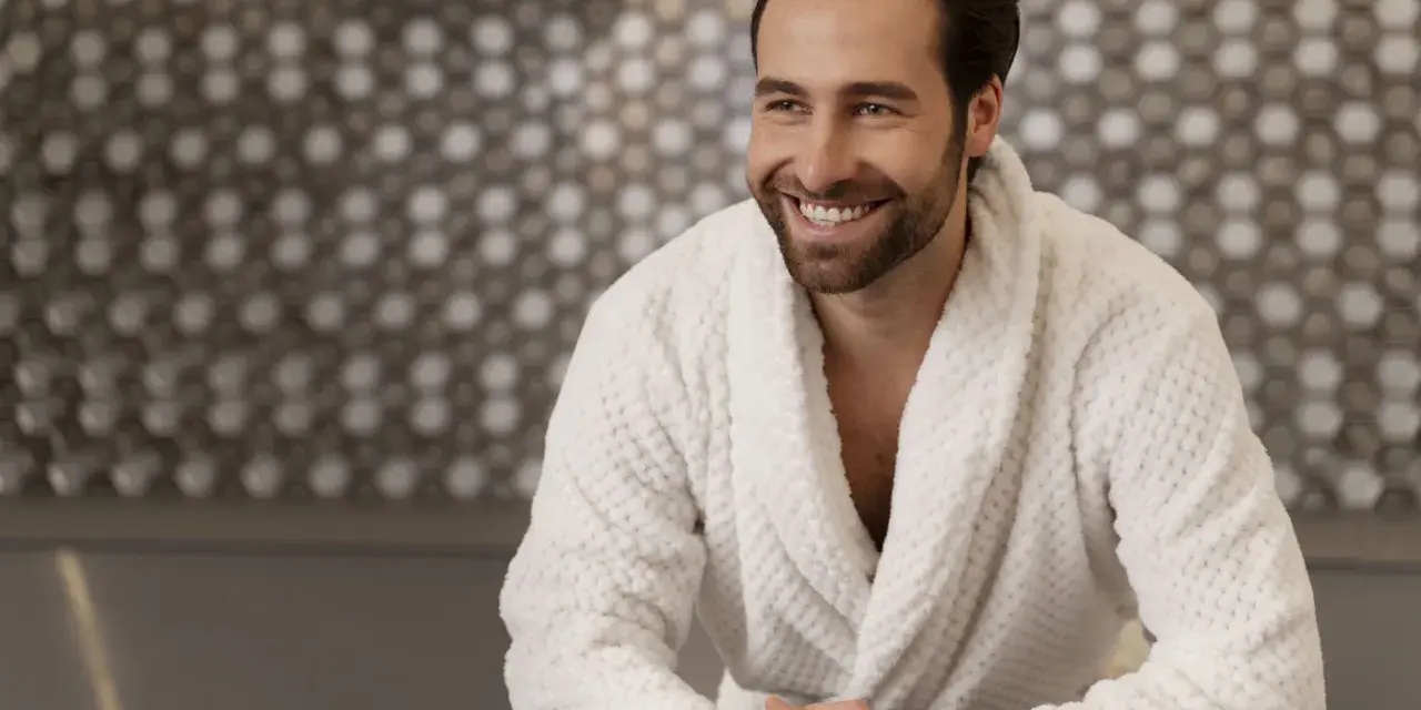 Uomo sorridente in un morbido accappatoio che asciuga bene, con uno sfondo a mosaico.