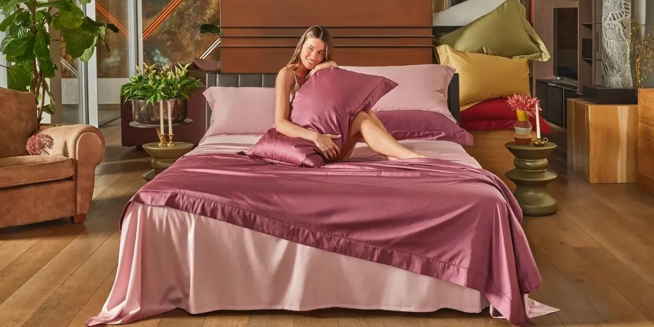 Donna sorridente su un letto con lenzuola rosa. Scopri come lavare le lenzuola nuove per mantenerle morbide e fresche.