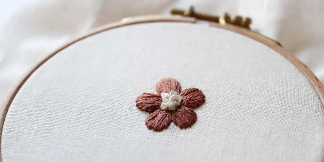 Piccolo fiore ricamato a punto pieno su tela grezza, con petali marroni e centro bianco.