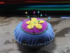 Puntaspilli fai da te a forma di fiore, con petali gialli su sfondo viola e base blu, decorato con cuciture a zig-zag verdi e gialle.
