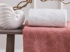 Asciugamani rosa e bianchi con motivi floreali in rilievo, disposti su un tavolino con una decorazione a conchiglia. La grammatura degli asciugamani suggerisce morbidezza e qualità.