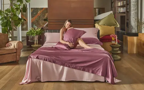 Donna sorridente su un letto con lenzuola rosa e copriletto prugna, che illustra la differenza tra piumone e trapunta.