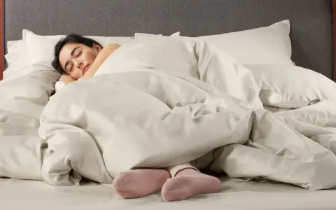Donna dorme avvolta in lenzuola in microfibra che sembrano farla sudare, con solo i piedi in calzini rosa visibili.