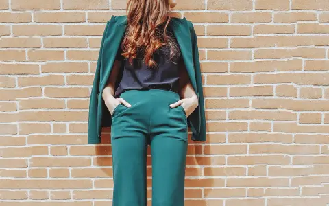 Donna con tailleur verde smeraldo, composto da giacca e pantaloni a palazzo, con le mani in tasca, appoggiata a un muro di mattoni.