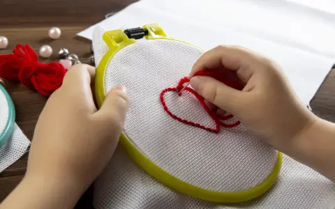Mani di bambino creano un cuore rosso con il punto erba a mano su tela bianca, circondato da perle e gomitoli di lana.