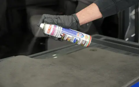Mano con guanto nero spruzza spray Idrostop su tessuto grigio per impermeabilizzare.