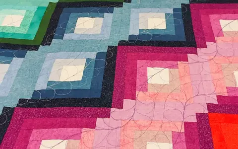 Dettaglio di una coperta realizzata con la tecnica patchwork, con blocchi a gradiente di colore dal blu al rosa, rifinita con cuciture decorative.