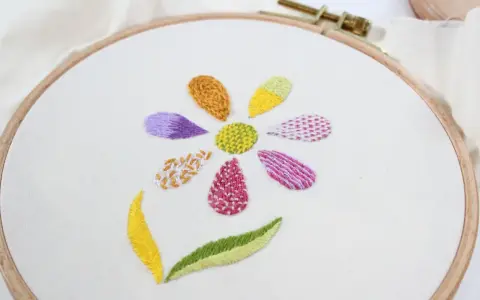 Fiore ricamato su tela bianca con petali colorati e diversi punti ricamo per riempire, che mostrano varie texture e tecniche.