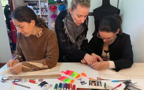 Tre donne si dedicano al cucito a mano, circondate da fili colorati, forbici e cartamodelli.