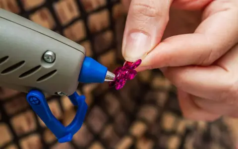 Applicazione strass su tessuto con colla a caldo. Una mano tiene un fiorellino fucsia mentre la pistola a caldo è pronta per incollare.
