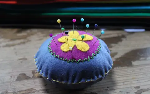 Puntaspilli fai da te a forma di fiore, con petali gialli su sfondo viola e base blu, decorato con cuciture a zig-zag verdi e gialle.