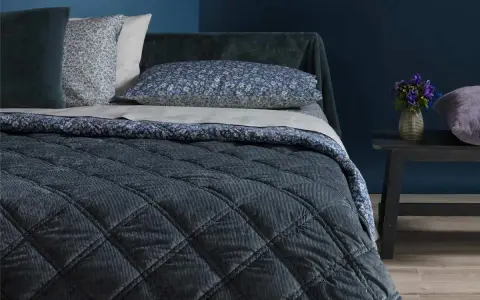 Letto con copriletto trapuntato blu scuro, cuscini floreali e un dettaglio che ricorda che il velluto si rovina con acqua.