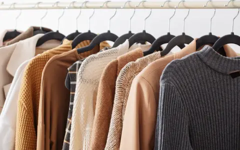 Una fila di capi d'abbigliamento in tonalità neutre, tra cui un maglione lavorato a maglia e un blazer in tessuto cupro lavaggio, pronti per essere indossati.