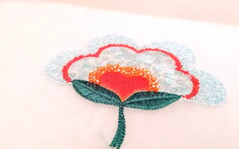 Un fiore stilizzato con petali azzurri e paillettes, un cuore rosso al centro e perline arancioni. Un'opera di ricamo francese che cattura la luce.