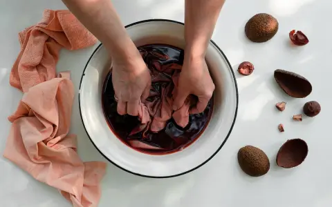 Mani che tingono un tessuto in una bacinella con un colorante naturale per tessuti. Avocado e bucce intorno.