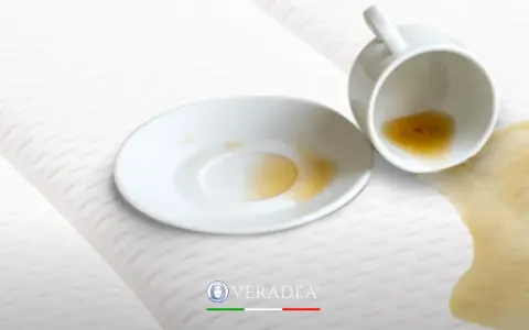 Tazza rovesciata su un materasso bianco, con liquido marrone versato. Scopri come disinfettare il materasso per eliminare macchie e odori.
