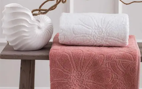 Asciugamani rosa e bianchi con motivi floreali in rilievo, disposti su un tavolino con una decorazione a conchiglia. La grammatura degli asciugamani suggerisce morbidezza e qualità.