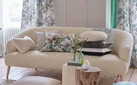 Divano in tessuto bouclé color crema, pronto per essere rifoderato, con cuscini decorativi e tende floreali sullo sfondo.