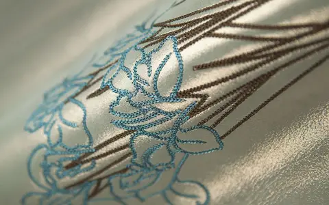 Dettaglio di un tessuto con ricamo floreale in punto pieno, con fili blu e marroni su sfondo cangiante.