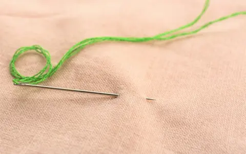Ago e filo verde pronti per ricamare nome a mano su stoffa color pesca.